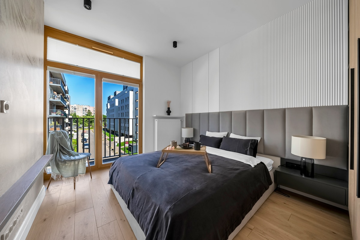 The modern bedroom features a spacious bed with dark bedding and accent pillows. Natural light enters through large windows, revealing a view of the exterior. Two bedside tables with lamps are positioned on either side of the bed, complemented by a cozy chair in the corner.