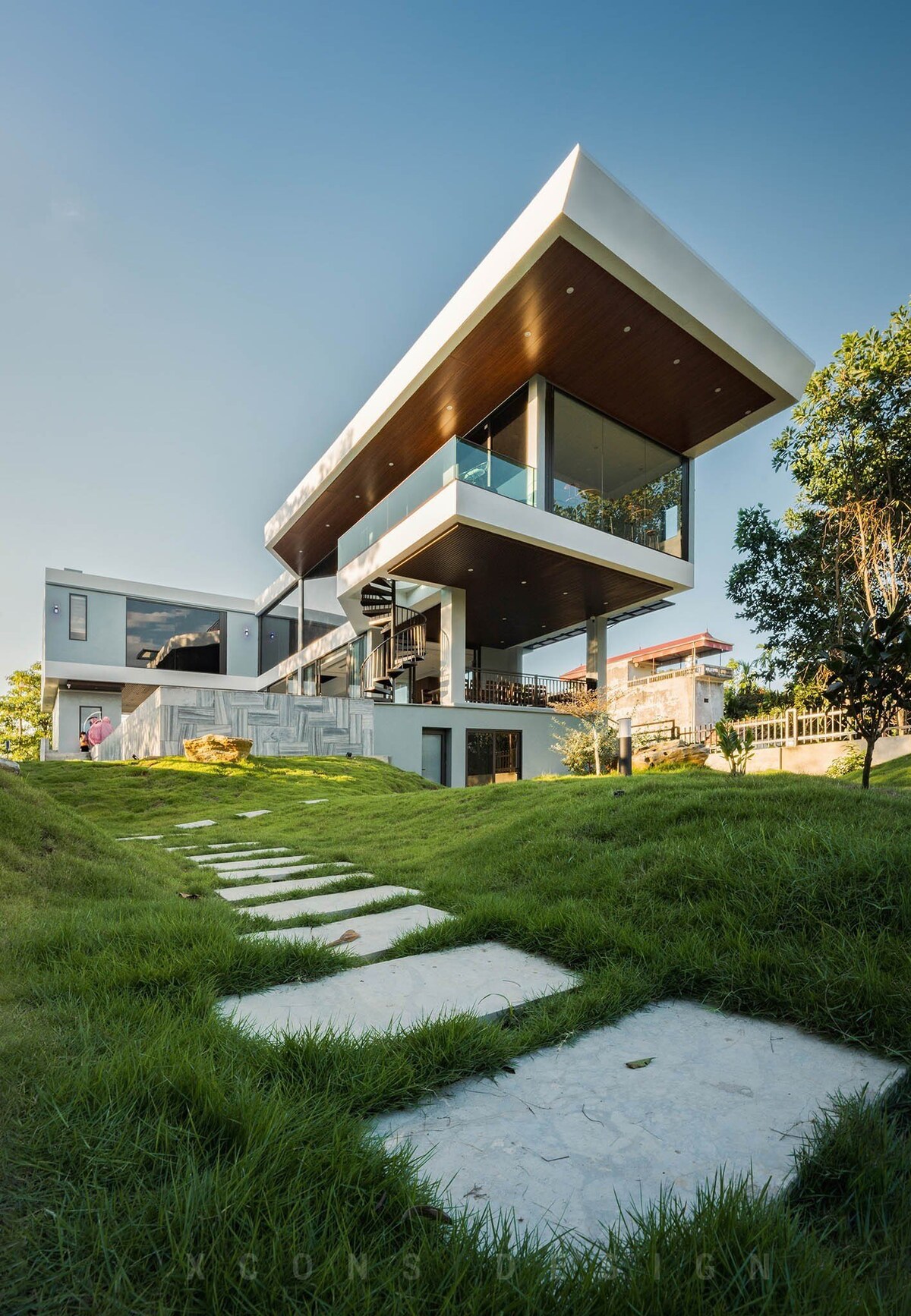 A modern two-story home is surrounded by lush green grass and stepping stones leading to the entrance. Large glass windows and an overhanging roof create a contemporary look. The structure features an open design that enhances the connection with the outdoors.