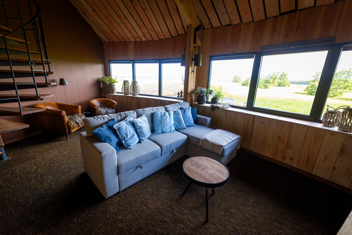 A cozy seating area features a light-colored sectional sofa adorned with blue cushions. Natural light floods in through large windows, offering views of the surrounding fields. The wooden accent walls and staircase add warmth to the space, complemented by a small round table.