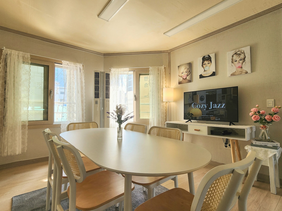 A dining area is shown with a large table surrounded by six chairs. Natural light is provided by multiple windows with sheer lace curtains. A wall-mounted television displays 'Cozy Jazz,' and decorative flowers adorn the table, creating a welcoming space for guests.