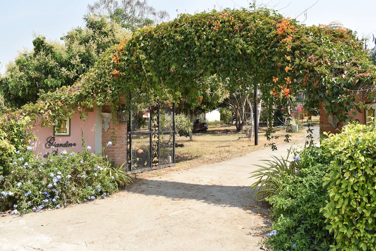 A charming entrance is framed by lush green vines and colorful flowers. A stone pathway leads through the archway, guiding guests toward a welcoming space. The surrounding area features well-maintained gardens with grass and various plants, creating a serene setting.