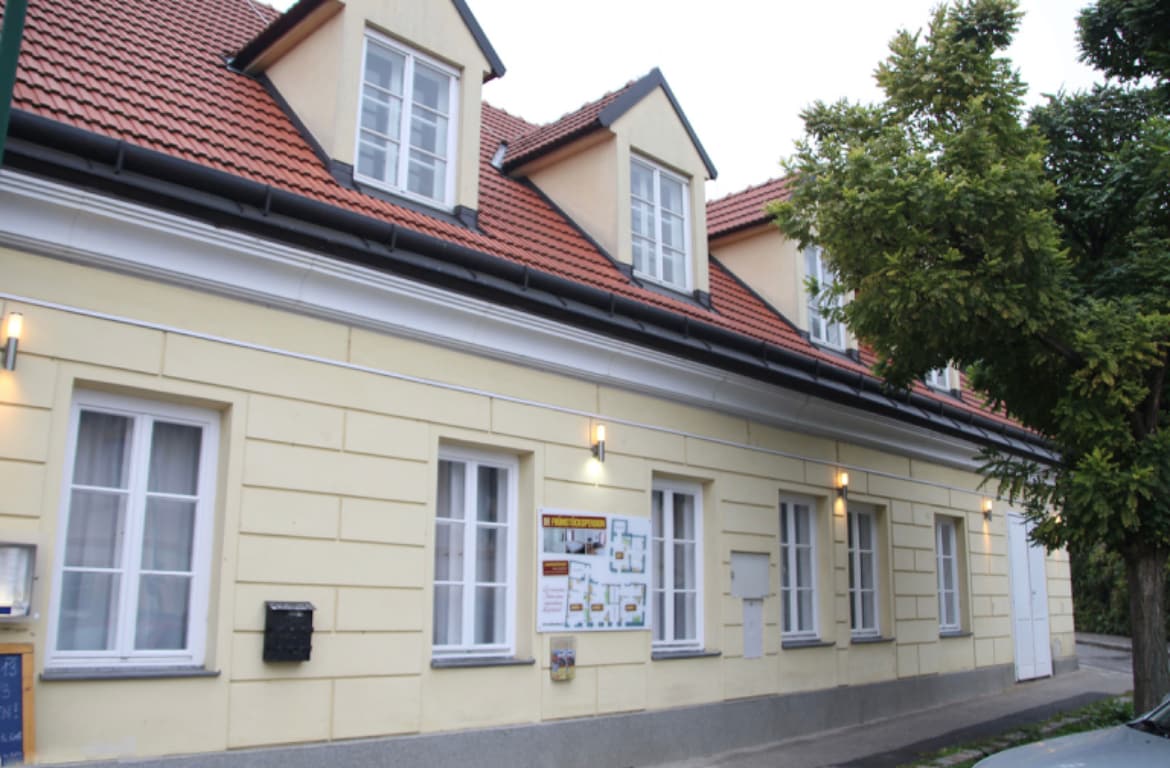 The exterior of a charming two-story house is shown, featuring light-colored walls and a red-tiled roof. Several windows are visible, providing natural light. A tree is located nearby, adding greenery to the setting. A wall-mounted information board enhances convenience for guests.