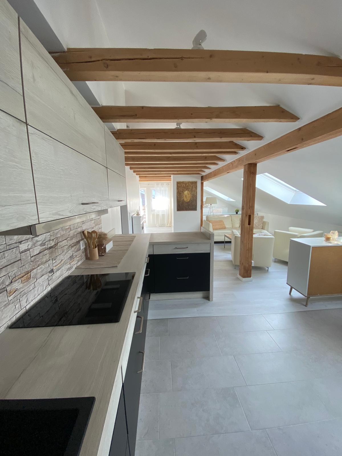 A modern kitchen is presented with light wood cabinets and sleek black accents. A stone wall adds texture, while exposed wooden beams lend character to the ceiling. Natural light fills the space, enhancing the open layout that connects to the living area.