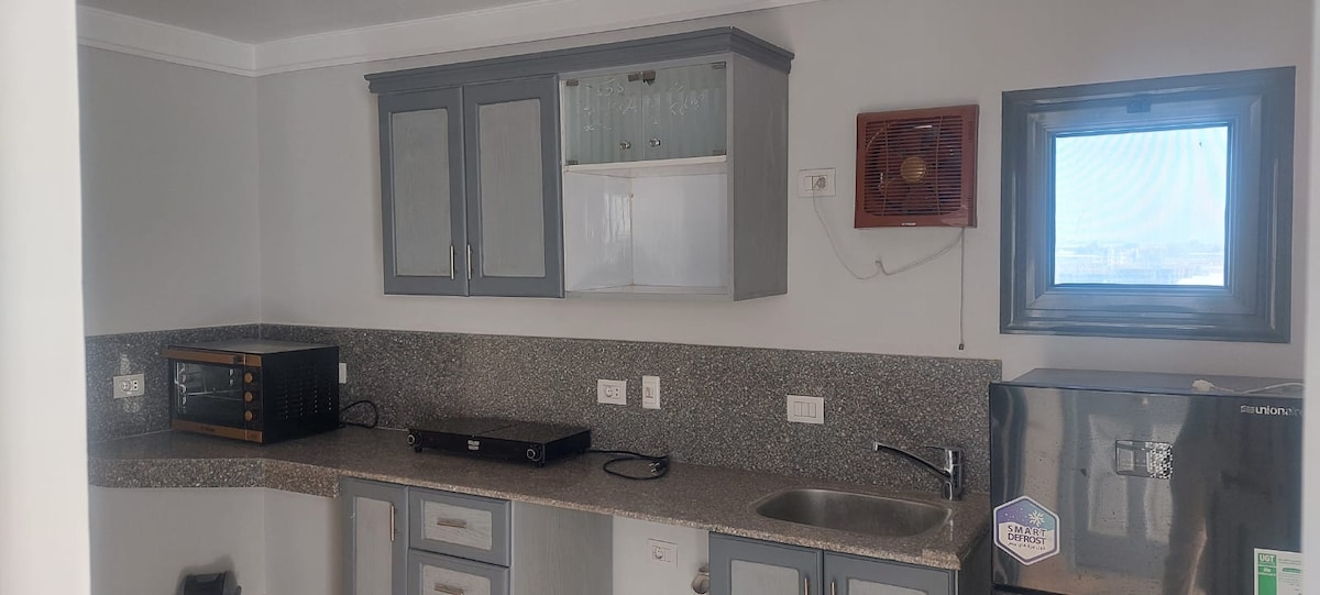 A functional kitchen area is equipped with essential appliances, including a microwave and a refrigerator. Grey cabinetry contrasts with light countertops, while a window allows natural light to brighten the space. A stove is also visible on the counter, providing convenient cooking options.