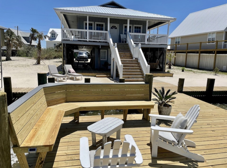 A spacious wooden dock features comfortable seating arrangements, including a built-in bench and two white Adirondack chairs. Steps lead to a charming two-story house with a wrap-around porch, while the surrounding sandy area is enhanced by palm trees and nearby homes.