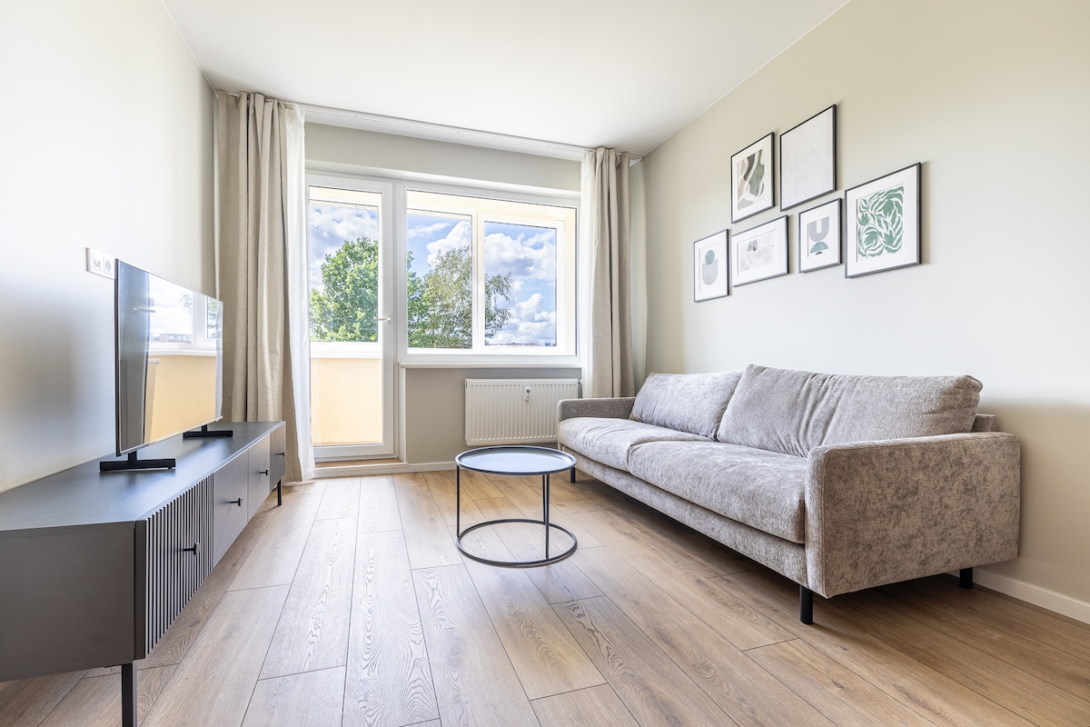 A living area features a comfortable sofa facing a modern flat-screen television. Natural light fills the space through a large window, adorned with light curtains. A round coffee table occupies the center of the room, complemented by framed artwork on the wall.