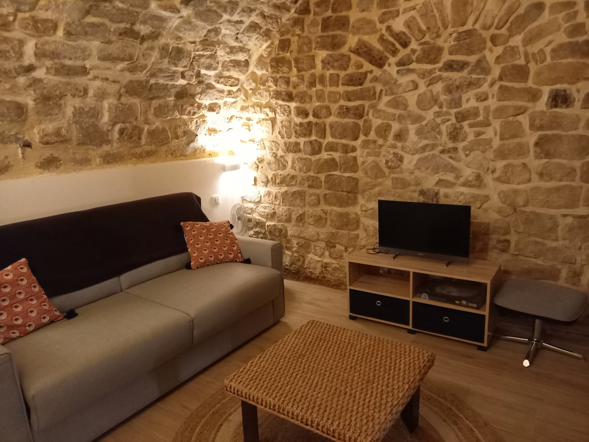 A cozy sitting area features a subdued grey couch adorned with two patterned cushions. A wooden coffee table sits in front, complementing the warm tones of the stone walls that lend character to the space. A flat-screen TV is mounted nearby on a wooden cabinet.