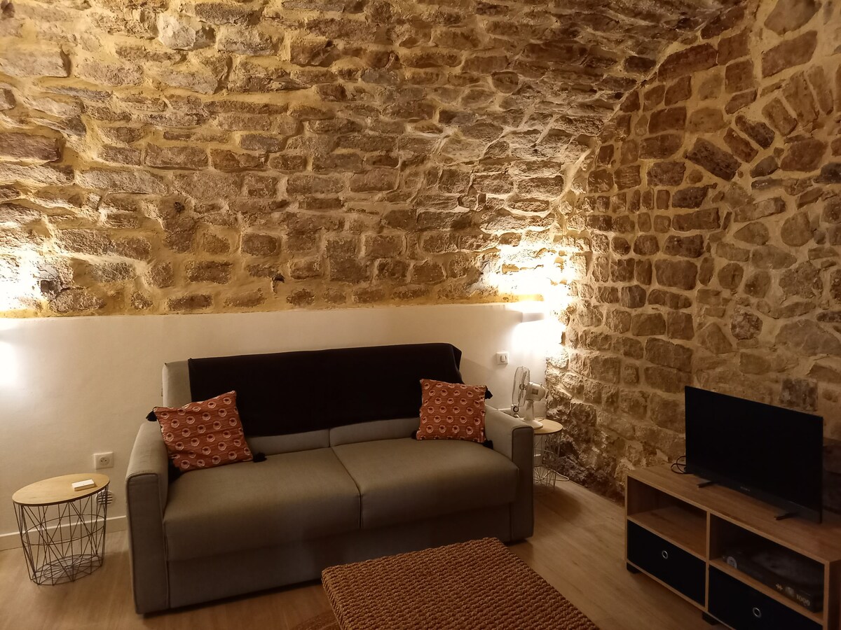 A cozy living area features exposed stone walls that add character. A gray sofa is complemented by decorative cushions, while warm lighting from adjacent fixtures enhances the ambiance. A wooden coffee table sits in front of the seating area, and a television is positioned on a media console.