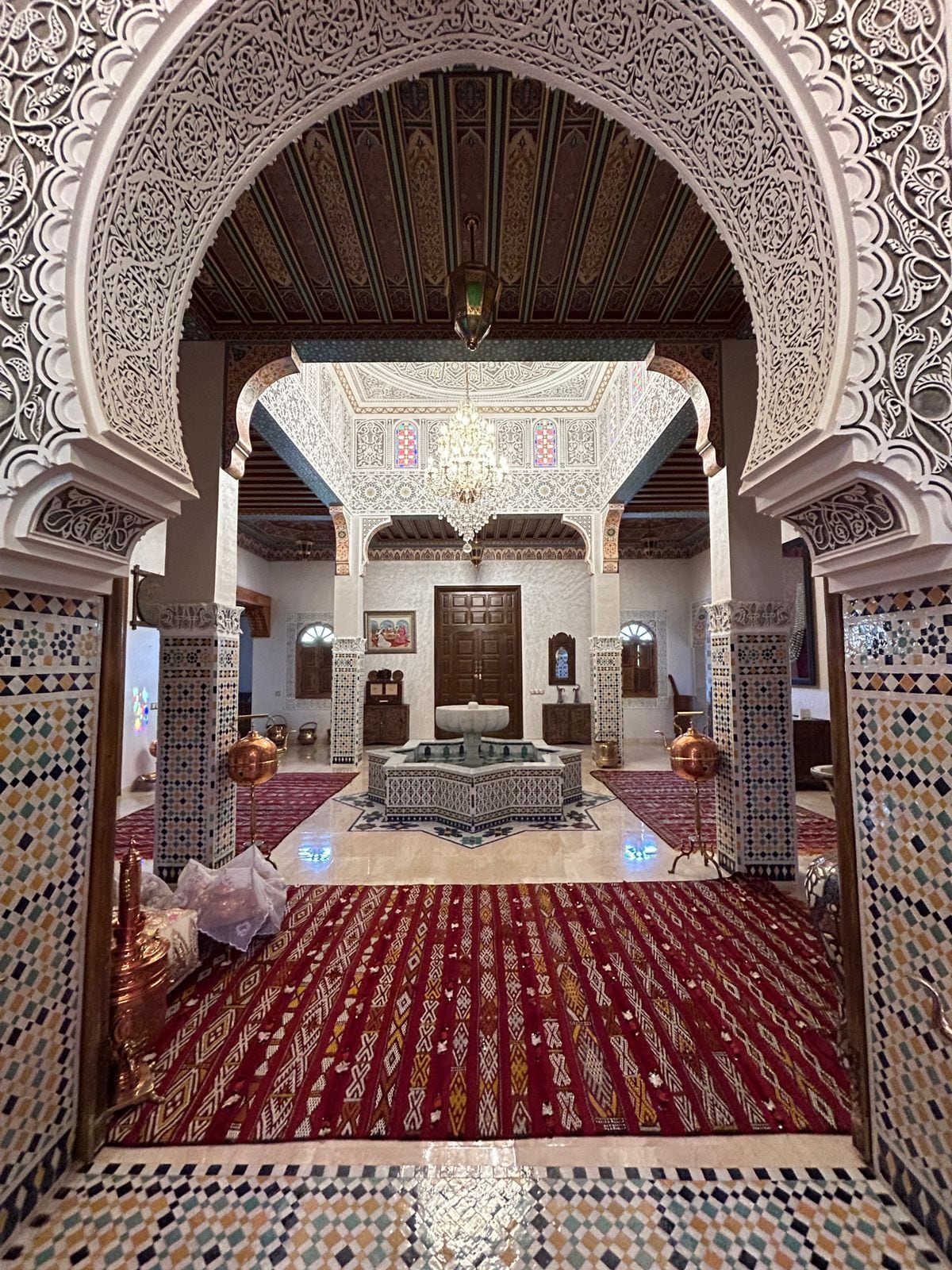 An intricately designed entrance leads into a spacious living area, featuring ornate arches and detailed tile work. A central fountain is surrounded by decorative furnishings. The rich, patterned carpet complements the warm lighting from a chandelier, creating a welcoming atmosphere.
