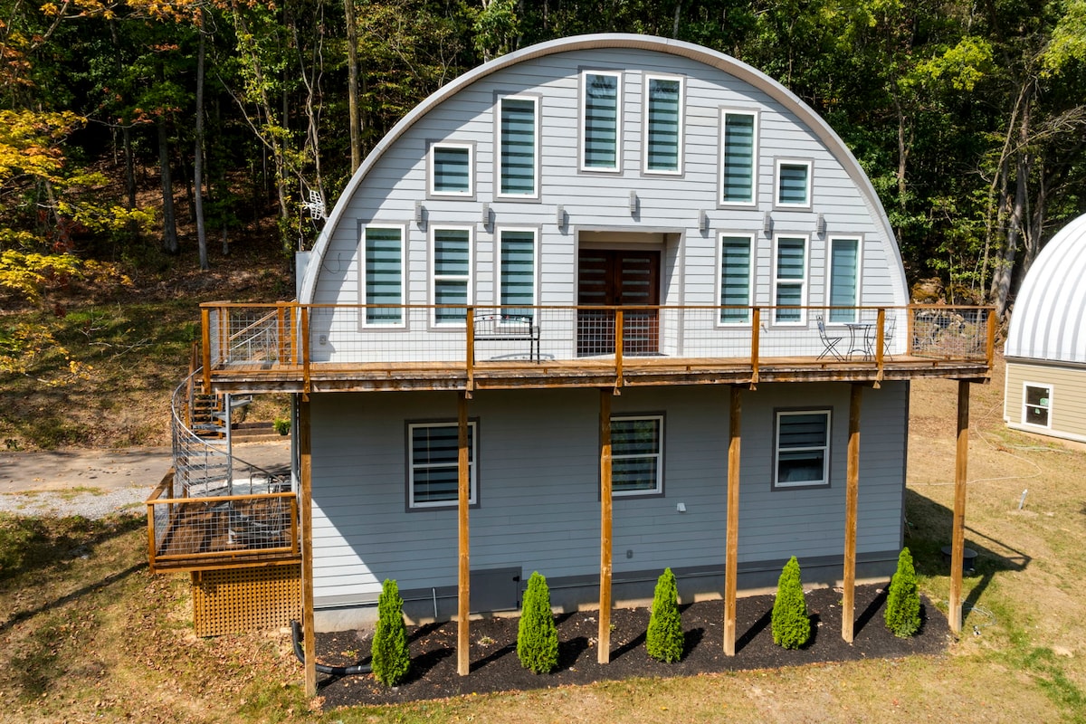 The exterior of the dome-style retreat is presented, showcasing a unique arched architecture with multiple windows. A spacious deck with wooden railings extends across the front, while neatly trimmed bushes line the base. Surrounding greenery enhances the serene atmosphere of the property.
