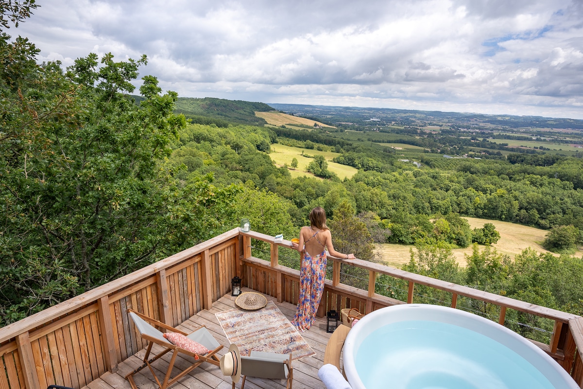 A private rooftop terrace is visible, featuring a hot tub and a wooden railing that offers a view of lush green hills. A person stands gazing at the landscape, surrounded by trees and open sky, creating a serene connection with nature.