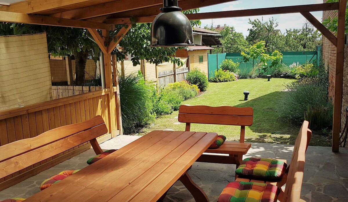 An outdoor seating area under a pergola is equipped with a large wooden table and benches, enhanced by colorful cushions. A spacious garden is visible in the background, featuring well-maintained greenery and trees, creating a relaxing environment for guests.
