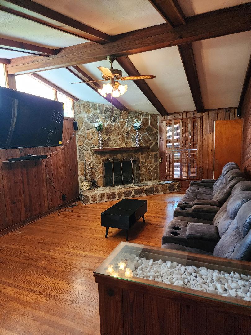 The living room features wooden walls and beams, creating a warm ambiance. A stone fireplace serves as a focal point, while a flat-screen TV is wall-mounted nearby. Comfortable seating is arranged around a small coffee table, and natural light filters through the windows with wooden shutters.