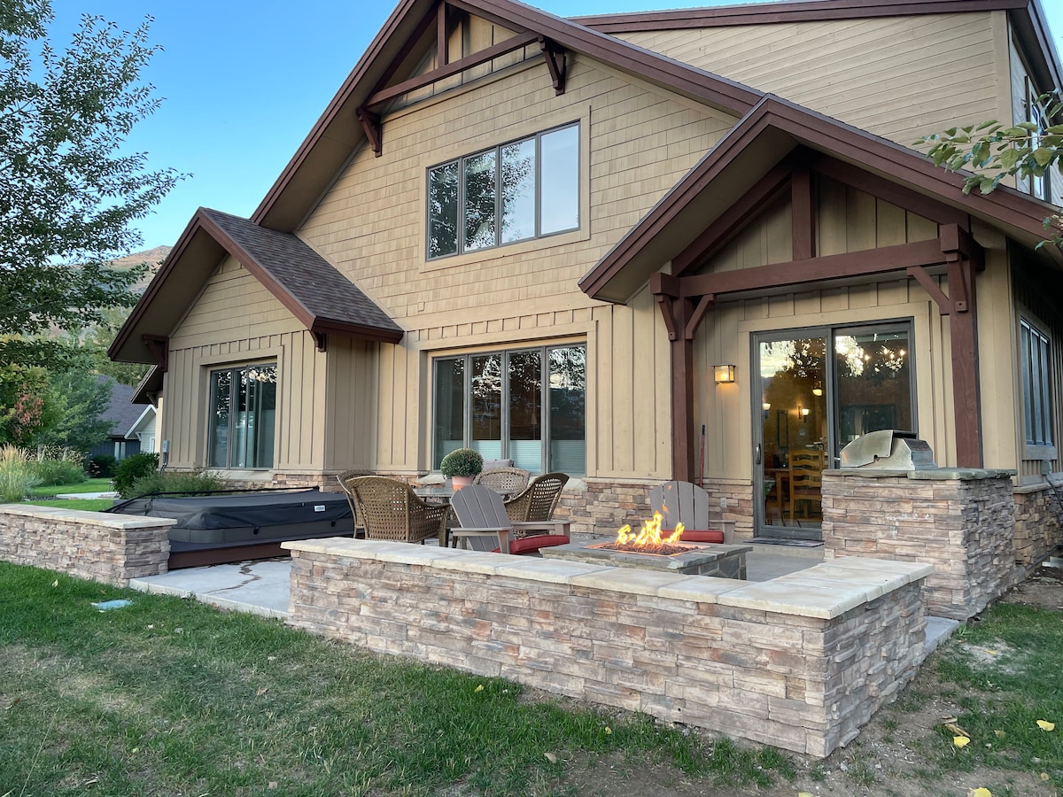 The exterior of the mountain retreat is showcased, featuring a stone firepit surrounded by outdoor seating and a private Jacuzzi. Large windows allow natural light to fill the inviting living space. Nearby, landscaped greenery enhances the tranquility of the setting.