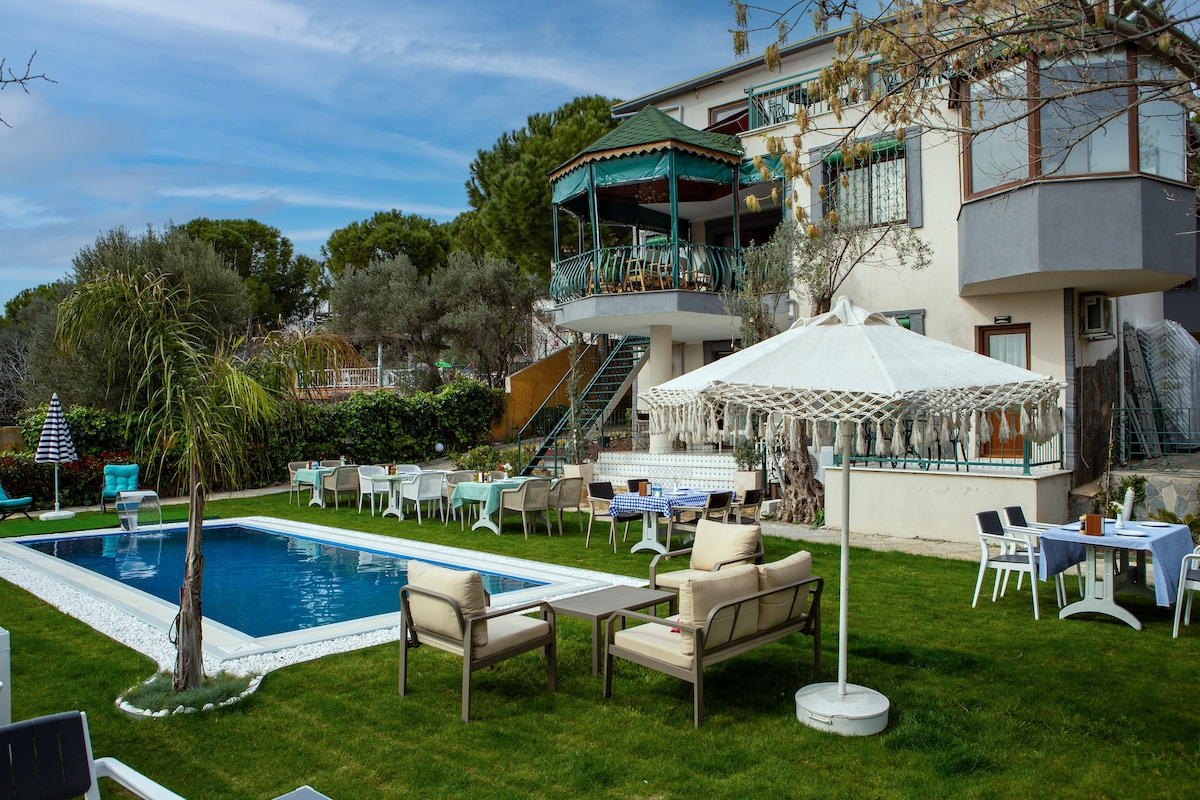 A well-maintained outdoor space features a rectangular swimming pool surrounded by manicured grass. Comfortable lounge chairs are positioned nearby, along with several dining tables. A large umbrella provides shade over a seating area, while a two-story building adds to the inviting atmosphere.