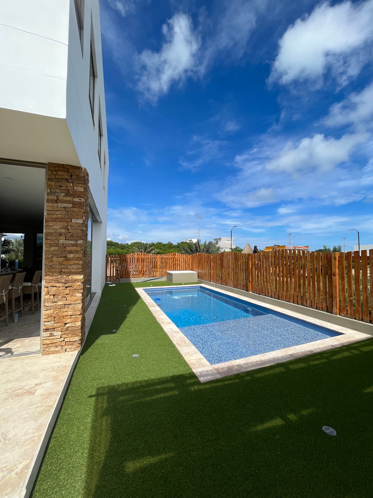 A private swimming pool is surrounded by a manicured grass area and enclosed by wooden fencing. Natural sunlight reflects off the water's surface under a bright blue sky, creating a serene outdoor space adjacent to the house.