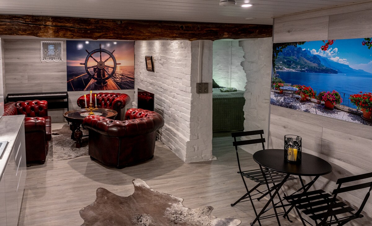 A cozy living area features a leather seating arrangement surrounding a circular table. Brightly colored wall art depicting landscapes enhances the ambiance. A stylish black and white rug is placed on the light wooden floor, while a small dining table set with chairs is visible in the corner.