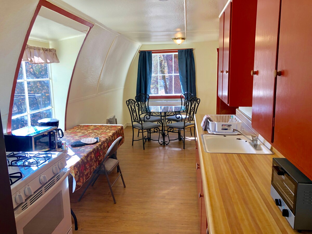 The kitchen and dining area is contained within an arched Quonset hut design. A round table is paired with four black metal chairs, and a floral tablecloth covers the table. Natural light fills the space through a large window with white curtains.
