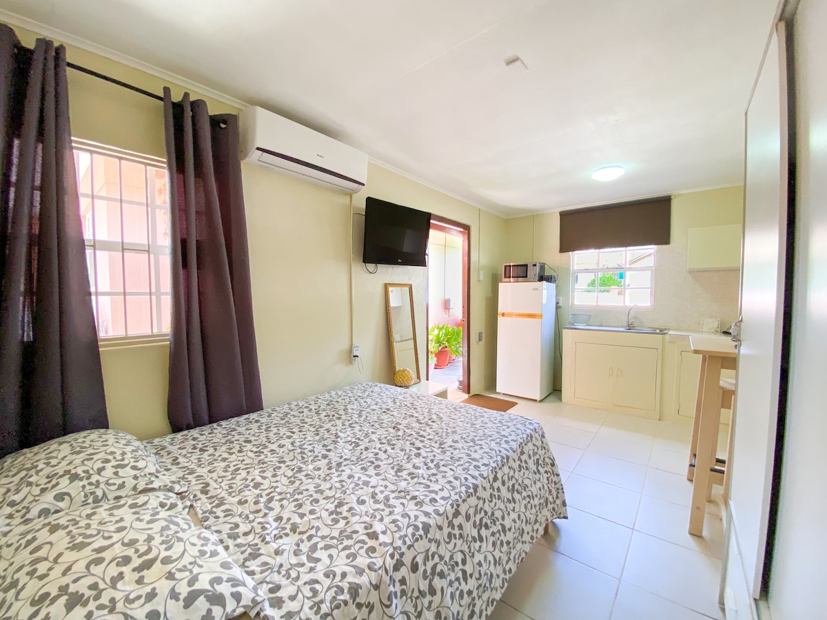 A cozy studio apartment is visible, featuring a spacious bed with a patterned bedspread. A kitchenette and refrigerator are located in the background, while large windows allow natural light to fill the room. Air conditioning and a television are mounted on the wall.