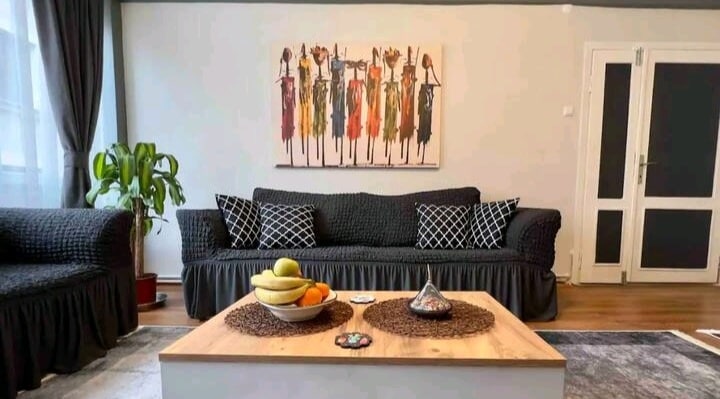 The living area features two spacious black sofas adorned with textured throw pillows. A wooden coffee table is positioned at the center, decorated with decorative items and fruit. A vibrant wall art piece depicting abstract figures adds character to the space, complemented by soft natural light filtering through large windows.