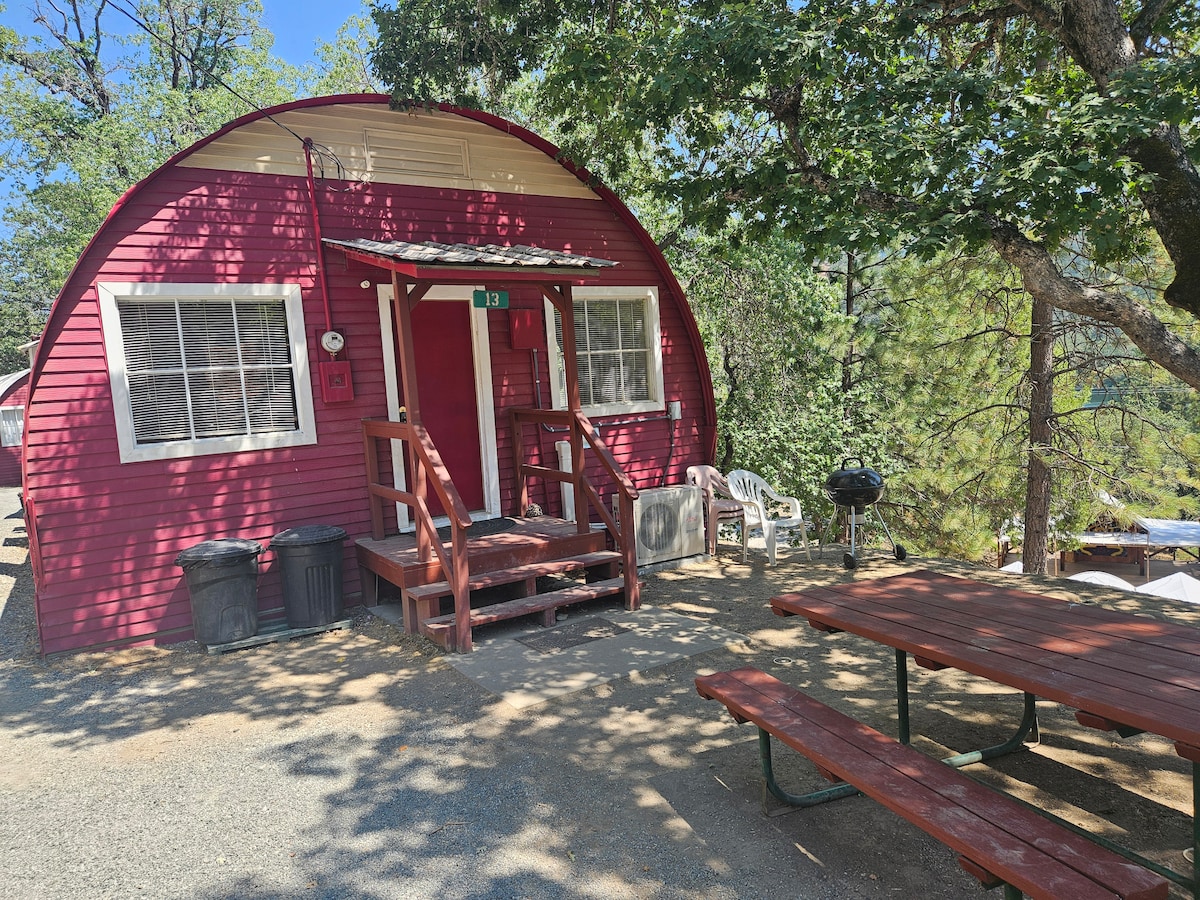 The exterior of a Quonset-style cabin is shown, featuring a bright red facade and a welcoming entrance with stairs. A picnic table and charcoal grill are positioned nearby under the shade of trees, with additional seating visible.