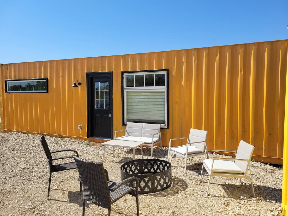 An outdoor seating area features a mix of chairs surrounding a firepit. The bright yellow shipping container serves as the backdrop, showcasing a black door and a large window. Gravel creates a natural ground surface, enhancing the inviting space for relaxation.