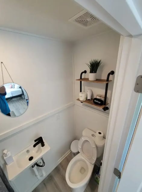 A small bathroom space features a compact toilet beside a wall-mounted sink. A round mirror is positioned above the sink, and a shelf above holds essential toiletries and a decorative plant. Neutral walls and flooring create a clean and simple environment.