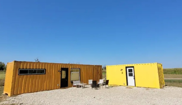 The Bumblebee Bunk House, constructed from yellow shipping containers, offers a unique exterior view. Outdoor seating is arranged in front, with a mix of chairs and tables positioned on gravel. A clear blue sky enhances the bright color of the structures.