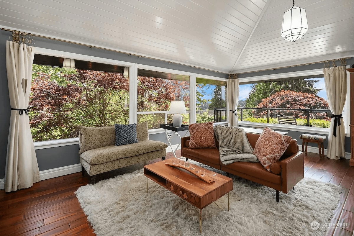 Antique-style main living room with natural light from south-facing windows, views of Mount Rainier and the water. Features vaulted ceilings, a large TV with audio system, indoor fireplace, ample seating, and hardwood floors.
