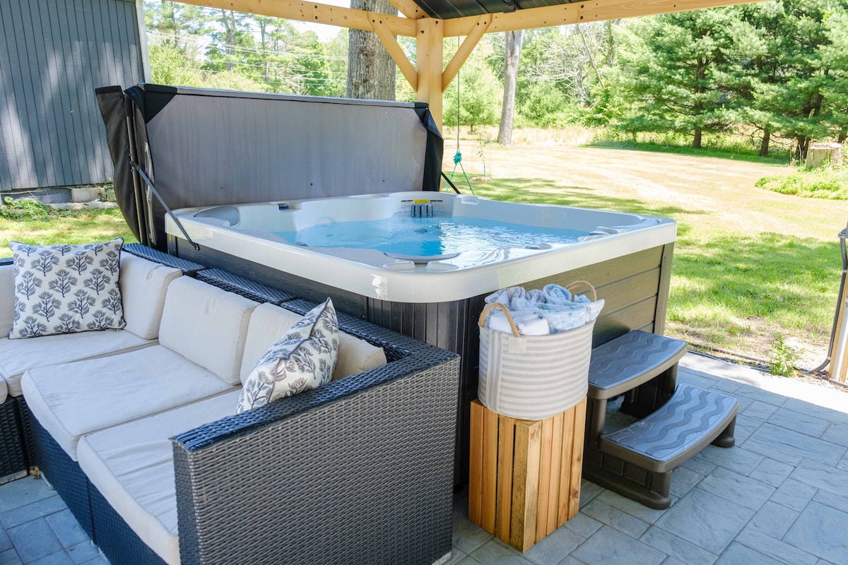 A hot tub is situated under a wooden gazebo, surrounded by a spacious outdoor area featuring greenery. A cozy seating area with patterned pillows complements the space. A small side table and steps lead to the hot tub, enhancing the inviting atmosphere.