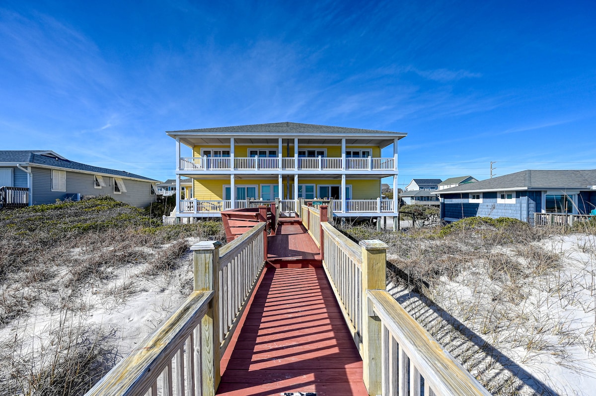 A two-story oceanfront house is showcased, with yellow siding and white railings. A wooden pathway leads from the sandy beach to the entrance, surrounded by natural grasses. Clear blue skies enhance the beachside setting, emphasizing the proximity to the ocean.