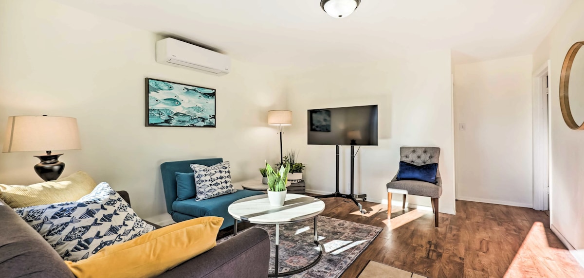 A bright living area features a mix of seating, including a blue sofa and an armchair. A coffee table is positioned at the center, with decorative plants nearby. Light streams in from a window, highlighting the neutral-toned walls and warm wood flooring.
