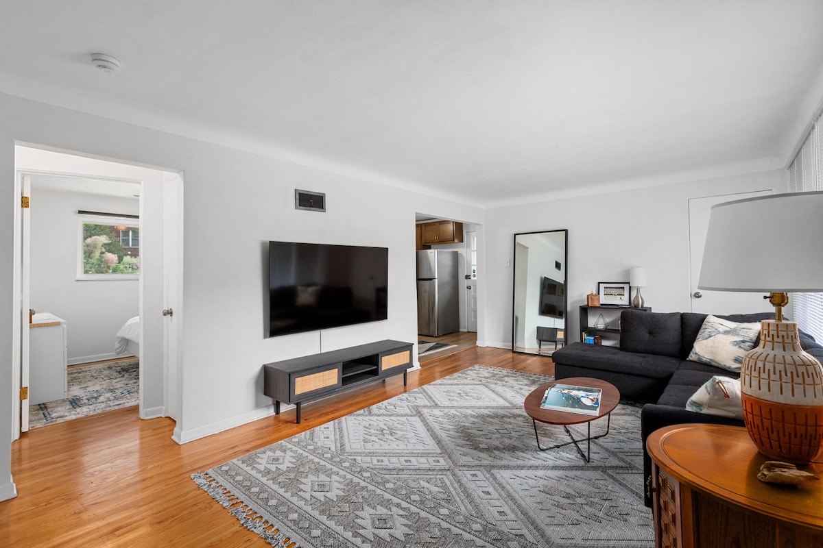 A spacious living area features a large flat-screen TV mounted on the wall. A dark sectional sofa is arranged around a low coffee table atop a decorative area rug. An entrance to the kitchen and another room is visible through a nearby doorway.