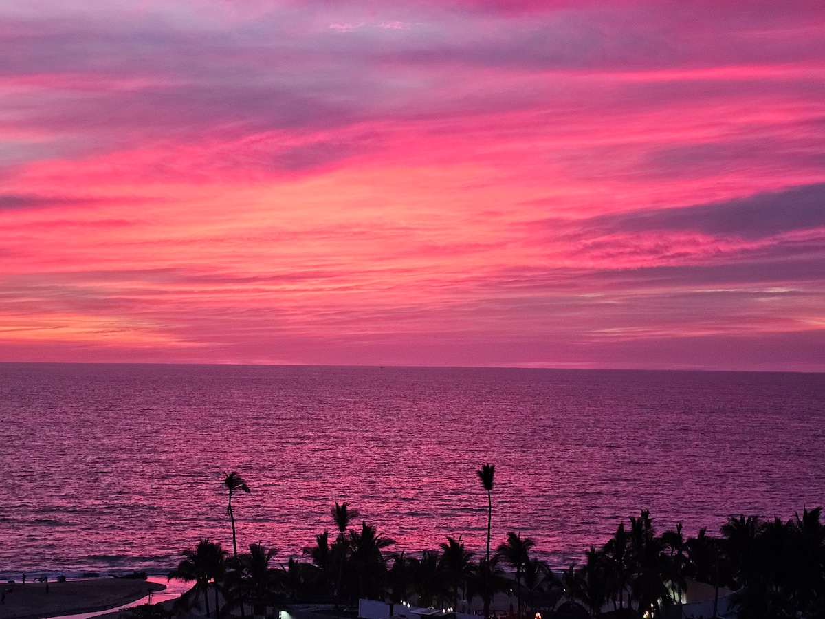 The image captures a vibrant sunset over the ocean, featuring shades of pink and purple in the sky. Silhouettes of palm trees line the foreground, framing the tranquil water while the coastline is faintly visible in the lower section.