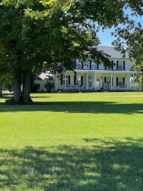 A well-maintained two-story farmhouse is set against a backdrop of lush grass and tall trees. A spacious porch features white railings and several decorative elements. The home is surrounded by an open area, creating a peaceful countryside atmosphere.