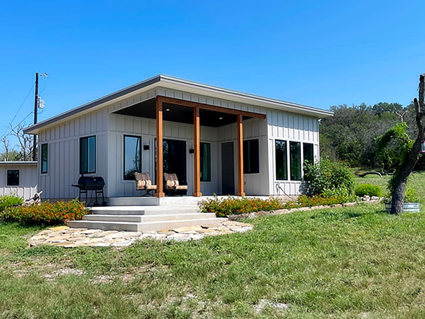 A modern cabin is featured with a light-colored exterior and large windows. A set of two chairs is placed on a covered porch, with a stone pathway leading to the entrance. Colorful flowers surround the area, and green grass extends into the distance.