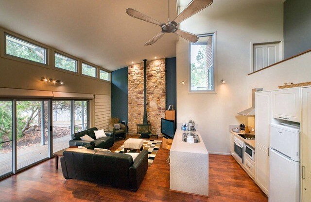 A spacious living area is illuminated by natural light from large windows. A stone fireplace acts as a central feature, while a sleek kitchen is visible, equipped with modern appliances. Comfortable seating options are arranged around a patterned rug, promoting a cozy atmosphere.