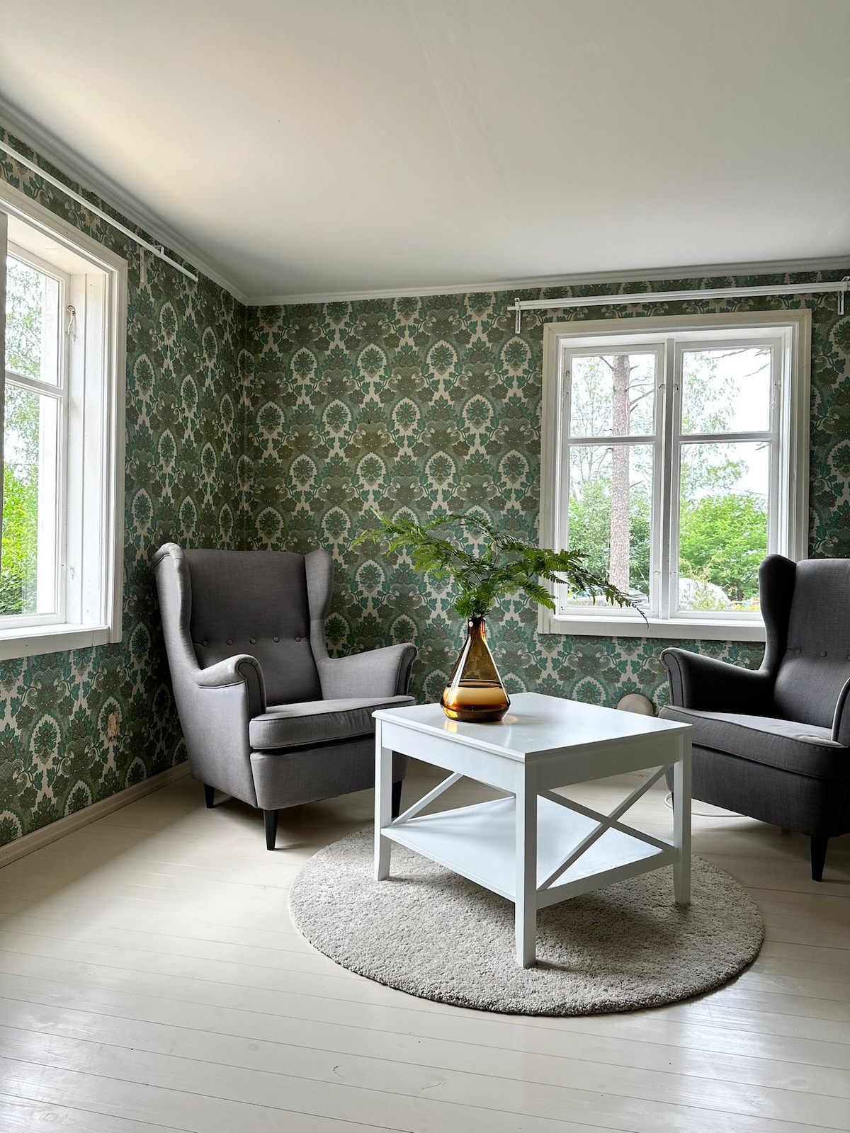 A light-filled seating area features two comfortable gray armchairs placed opposite each other. A simple white coffee table rests on a soft round rug, while large windows invite natural light and offer views of the surrounding greenery.