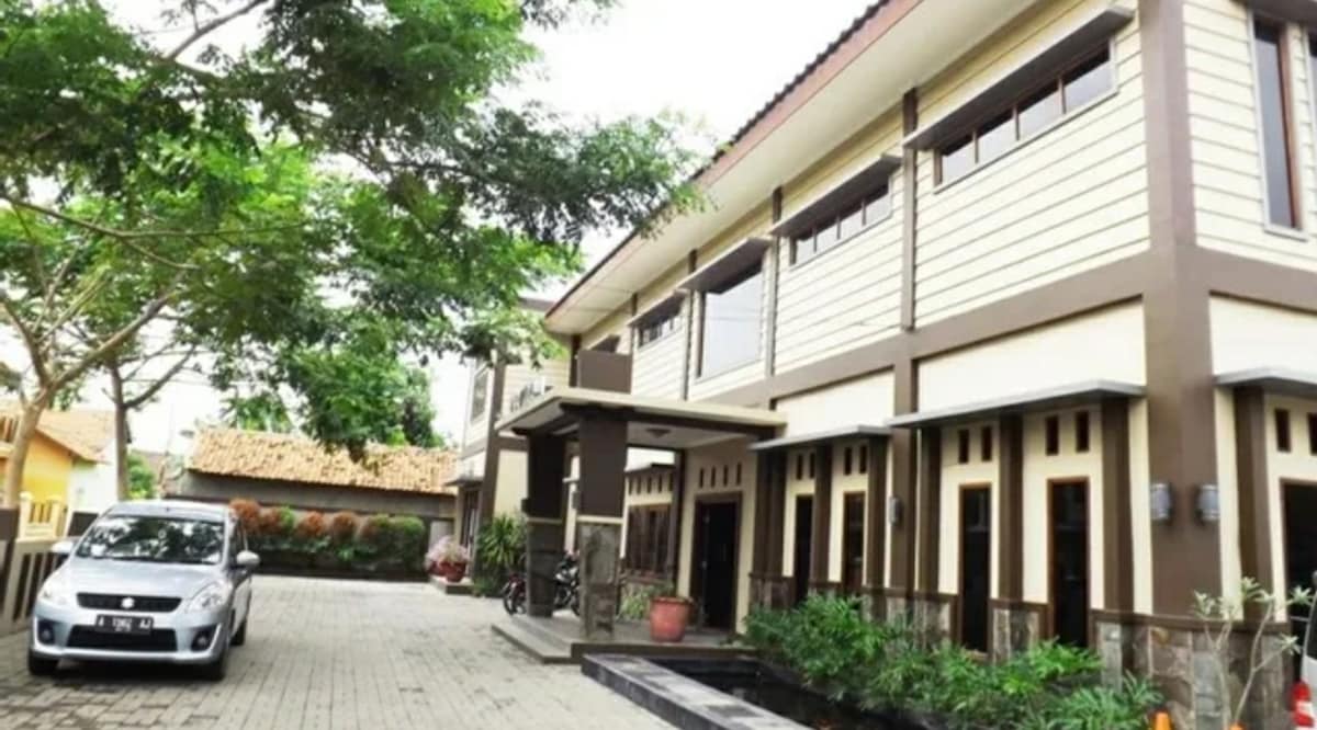 The exterior view of a modern multi-story building is shown, featuring a covered entrance with a clean, paved pathway. Lush greenery surrounds the property, and a parked car is visible in the driveway. Large windows allow natural light to illuminate the interior.