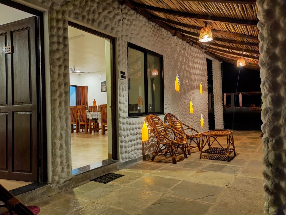 An outdoor patio area is featured, showcasing a textured stone wall and traditional bamboo roof. Two woven chairs and a low table sit on the stone floor, while lanterns hang softly illuminated around the space, inviting a warm ambiance for relaxation.