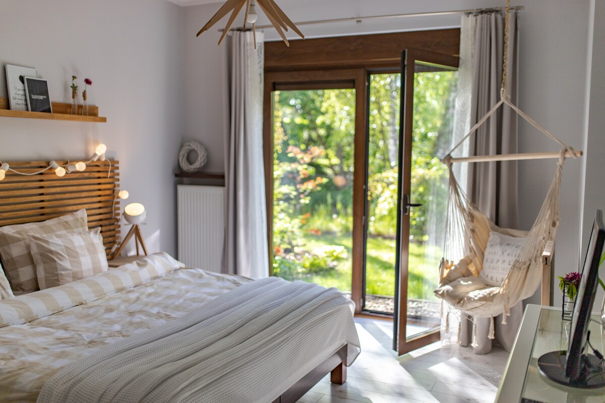 A serene bedroom is featured, highlighting a spacious bed with a neutral striped coverlet. Light streams through the large open doors, leading to a green garden area. A cozy hanging chair adds to the inviting decor, along with soft lighting from the bedside lamp.