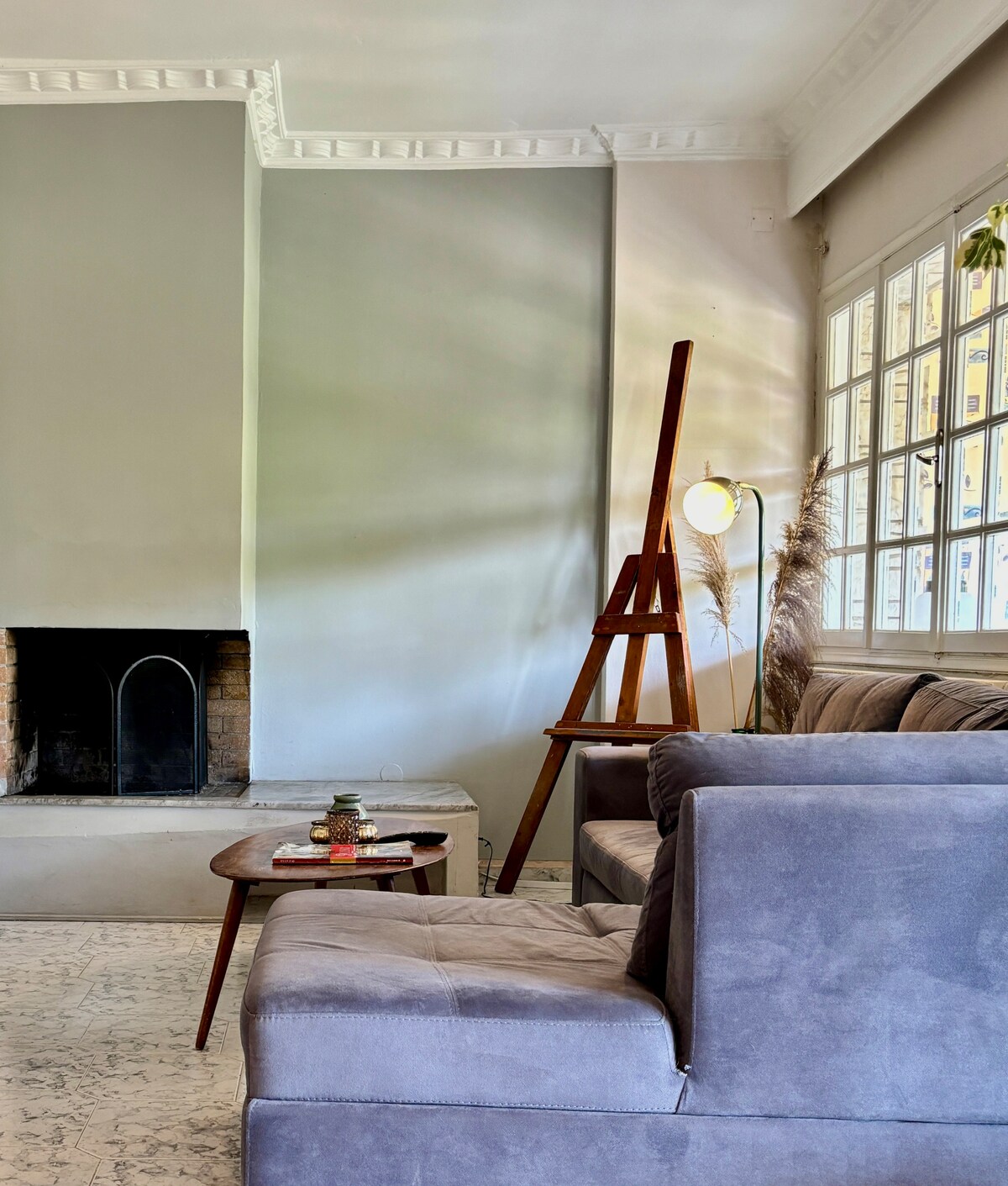 A cozy living space features a comfortable grey sofa paired with a low wooden coffee table. A simple fireplace is positioned on one side, while a wooden easel and floor lamp are placed to create a functional area. Bright natural light filters through large windows.
