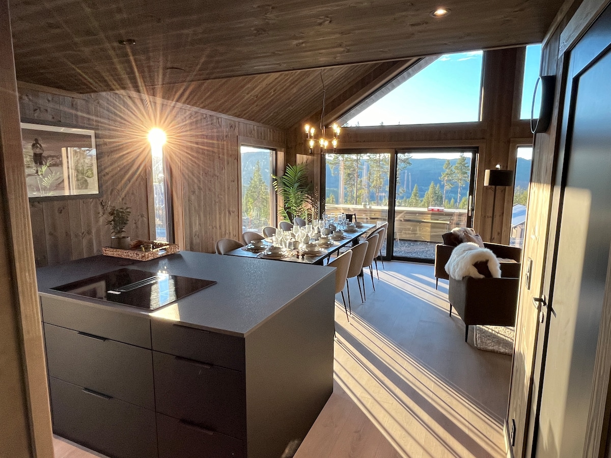 A modern kitchen and dining area features a sleek island and a long dining table set for a meal. Large windows allow natural light to illuminate the space, highlighting wooden accents and gentle outdoor views. A cozy seating arrangement is positioned nearby.