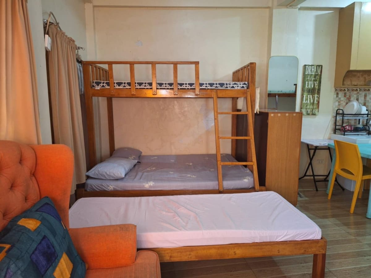 A bedroom area features a wooden bunk bed with a top and a lower twin-sized mattress. A simple wooden dresser stands nearby. A single bed with a light-colored mattress is positioned beside an orange couch adorned with a colorful cushion. Natural light filters in through window coverings.