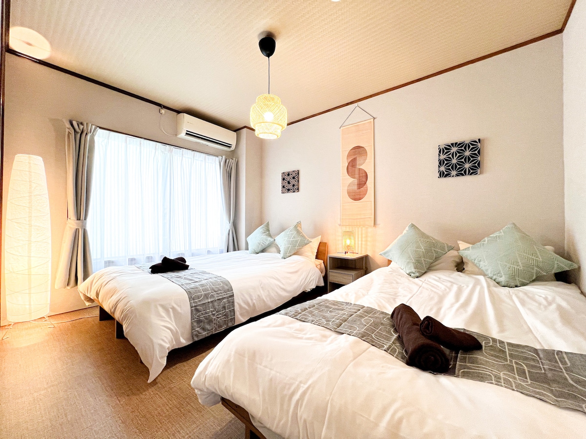 The bedroom features two spacious beds with soft white bedding and decorative cushions. Natural light is welcomed through a large window, complemented by sheer curtains. A minimalist floor lamp and wall art add subtle character to the serene space.