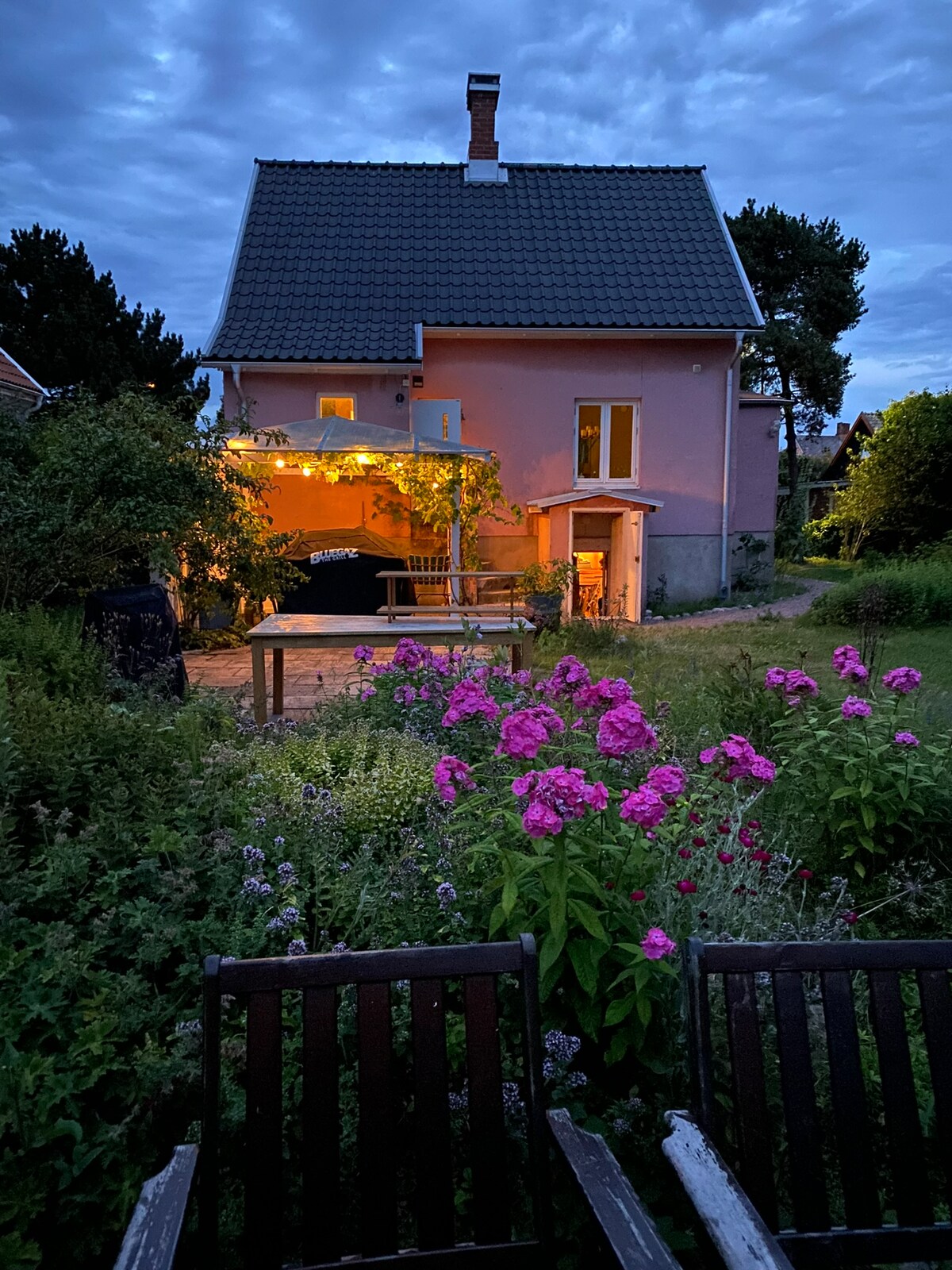 A charming pink house is illuminated under an evening sky, with soft lights glowing around a covered outdoor seating area. Lush plants and vibrant flowers create a colorful garden surrounding a wooden table and benches, inviting relaxation and outdoor dining.