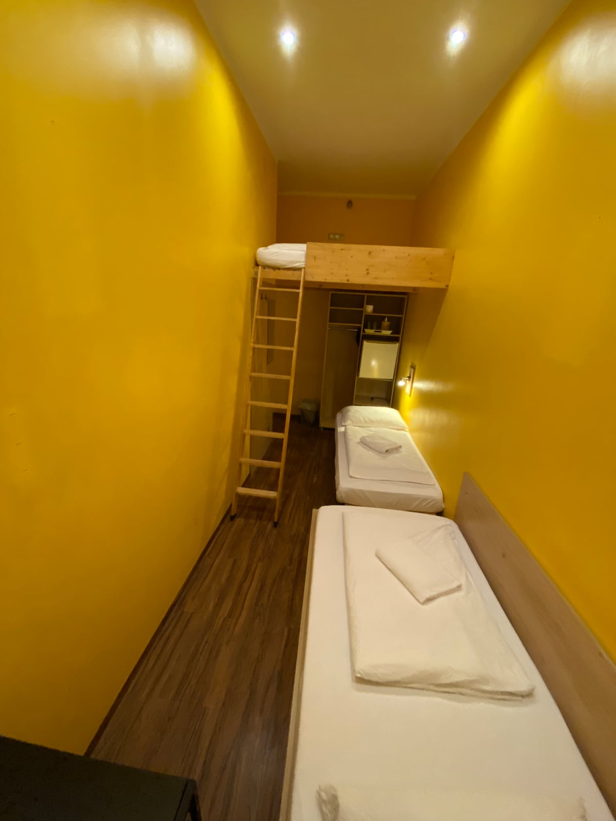 A narrow room features vibrant yellow walls, enhancing the bright ambiance. A single bed is positioned beneath a lofted double bed. A wooden ladder provides access to the upper sleeping area. Soft lighting illuminates the space, while wooden flooring enhances the overall warmth.