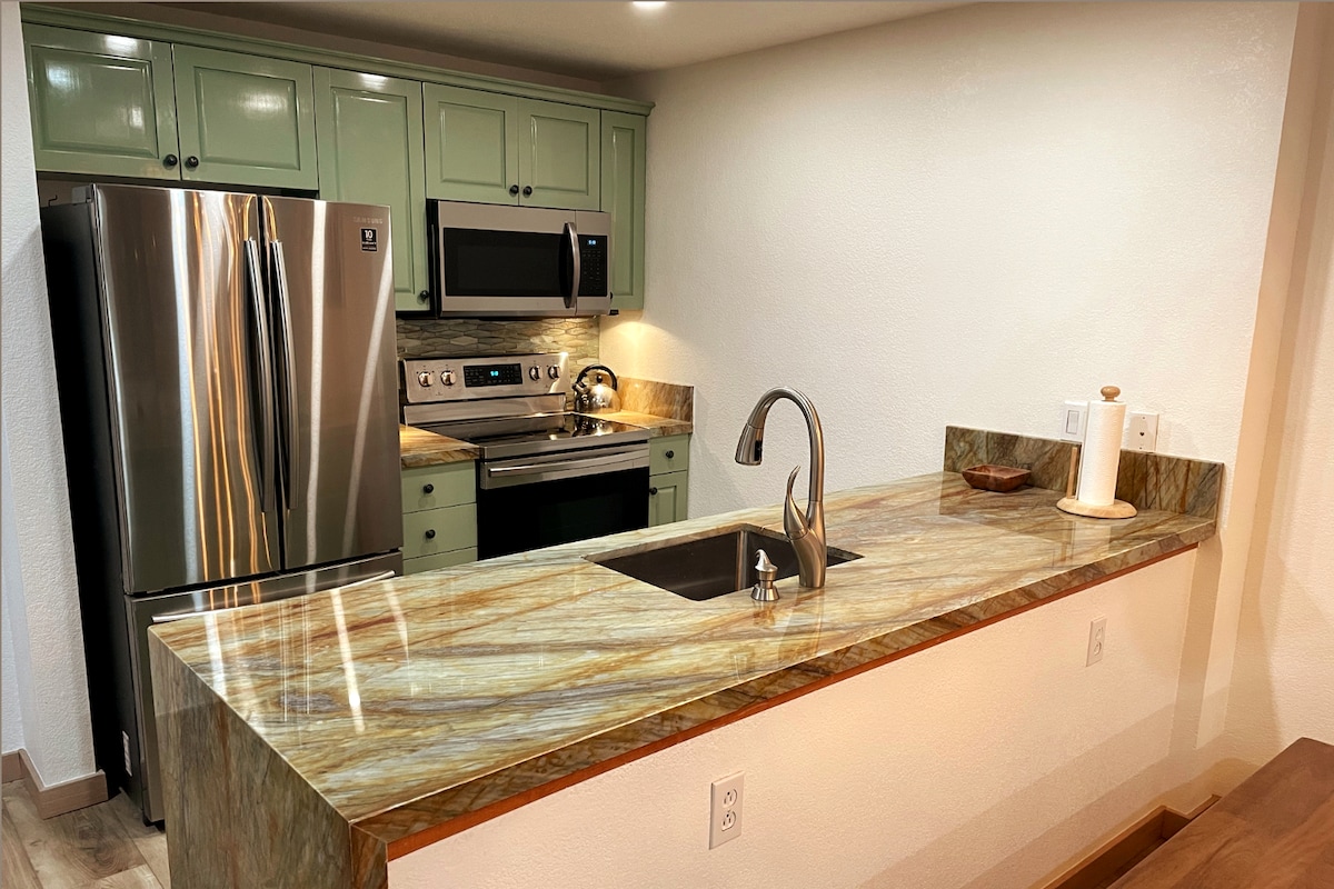 The kitchen features green cabinetry and modern stainless steel appliances, including a full-size refrigerator and microwave. A striking granite countertop extends over the bar area, with a sleek sink integrated into the design, providing functional elegance for meal preparation.