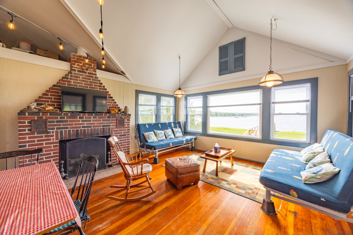 A spacious living area is highlighted by a brick fireplace and large windows offering views of the river. Comfortable seating includes a blue futon and wooden rocking chair. A wooden coffee table is centered on the hardwood floor, with a dining table nearby adorned with a red checked tablecloth.