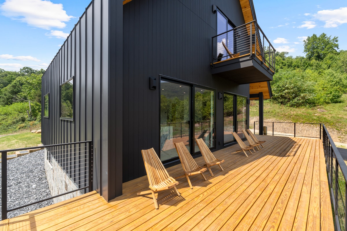 A spacious wooden deck extends from the modern black-clad exterior of the home. Five wooden lounge chairs are arranged for relaxation, overlooking the greenery surrounding the property. Large glass windows provide views of the interior and the natural landscape beyond.